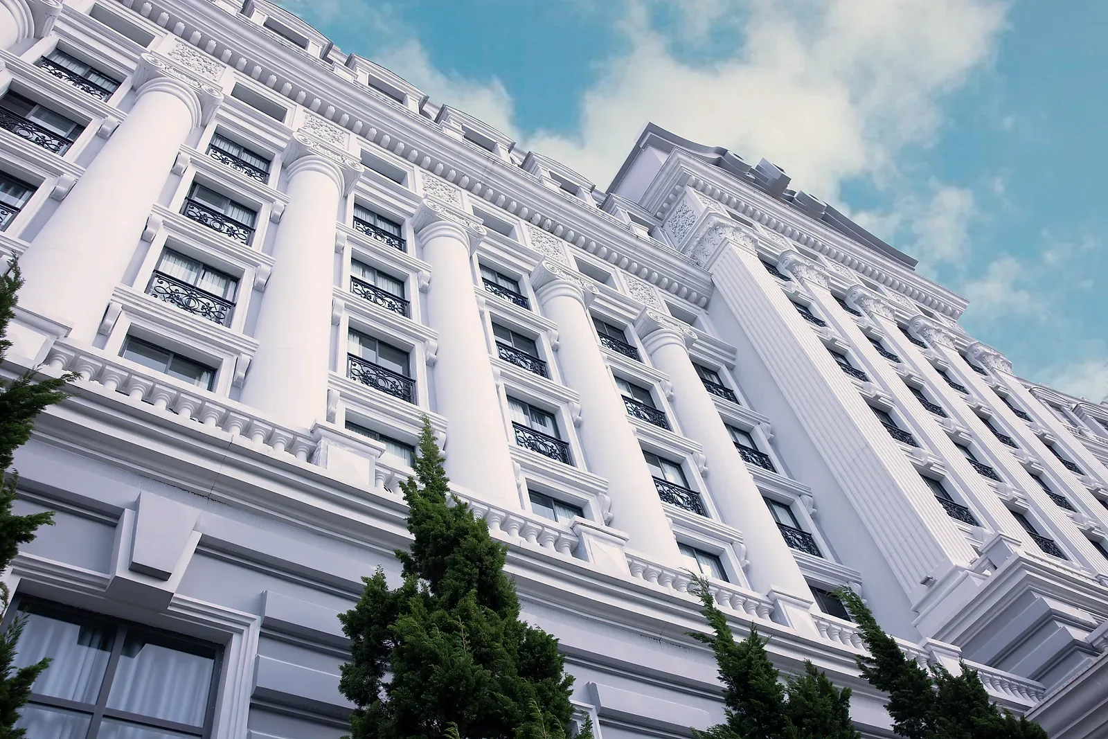 Mặt tiền trắng của MerPerle Dalat Hotel với hàng cột lớn và ban công sắt, nhìn từ dưới lên giữa rặng thông