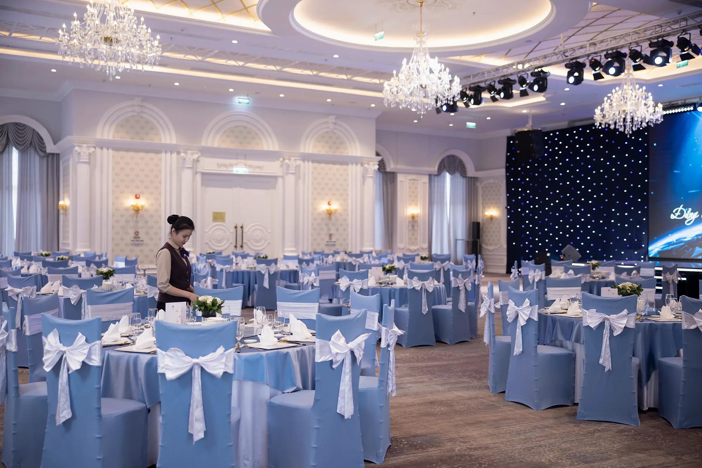Một người đang chuẩn bị bàn tiệc trong sảnh Romance bố trí bàn tròn xanh trắng.