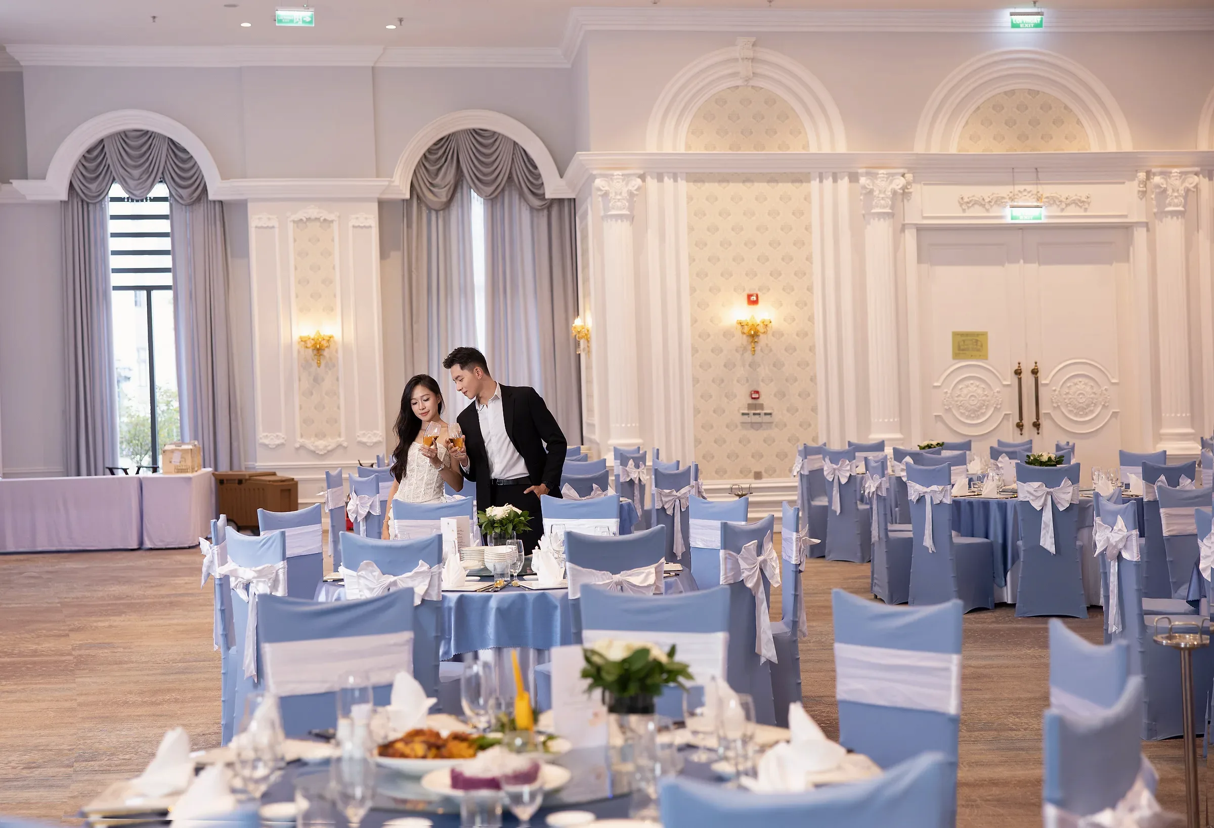 Hai người cầm ly bên bàn tiệc tròn tông xanh trắng trong sảnh Romance.