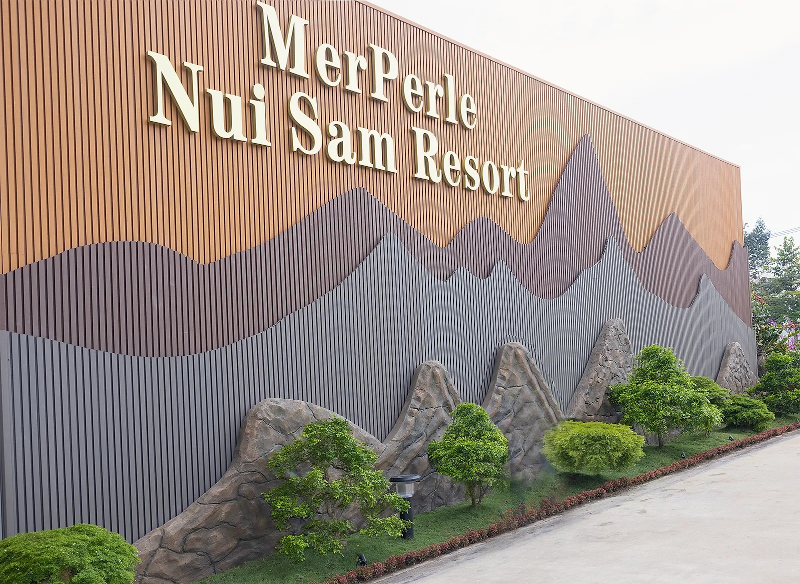 Tường tên MerPerle Nui Sam Resort ốp nan đất nung, bên dưới là phù điêu những lớp núi và tượng đá